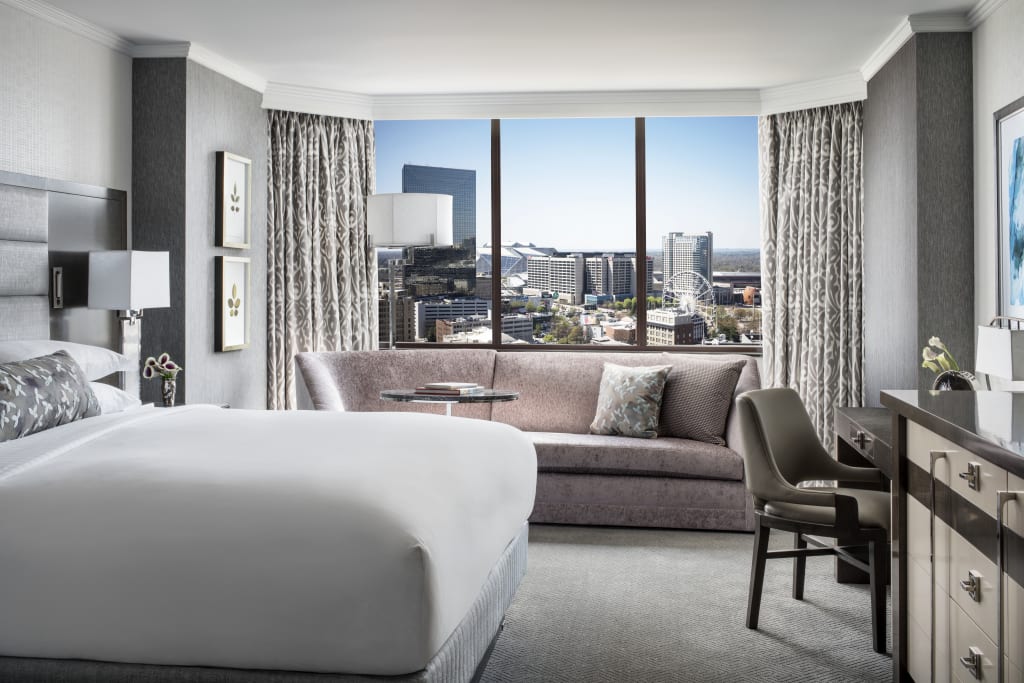 A modern hotel room with a king bed, sofa, desk, and large windows overlooking a cityscape in Atlanta, Georgia.