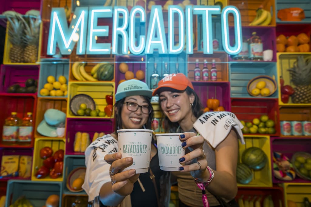 Two women cheerfully hold up cups of tequila in front of a colorful fruit display in Atlanta, Georgia.