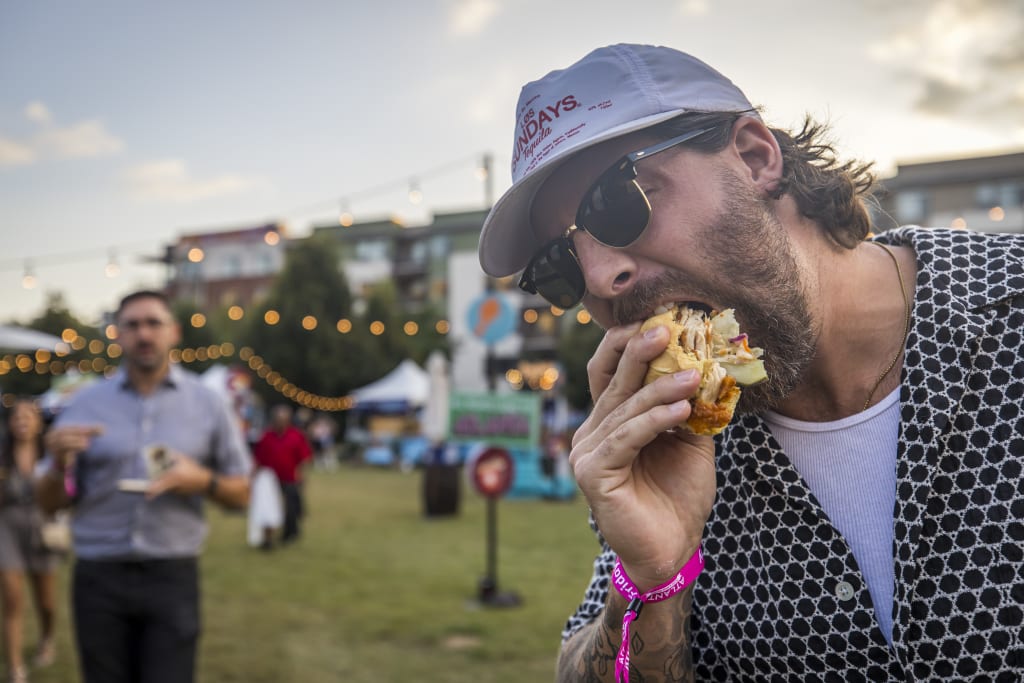 A man in a black and white patterned shirt and cap eats a sandwich at an outdoor event in Atlanta, Georgia.