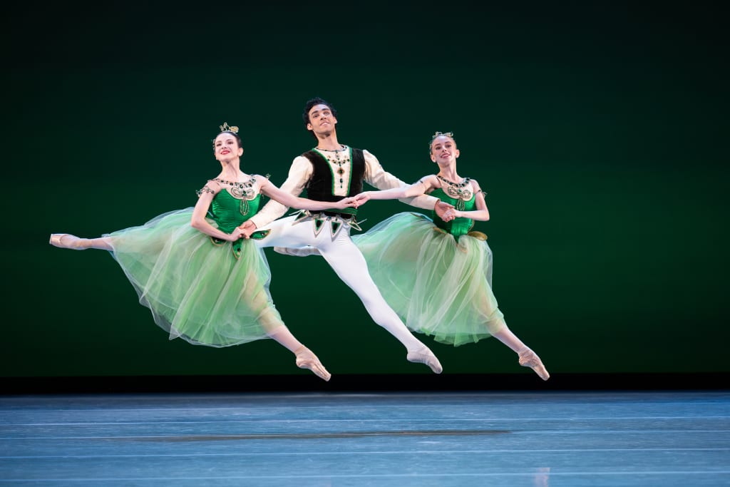 Three ballet dancers in green tutus and crowns perform a synchronized leap on stage in Atlanta, Georgia.