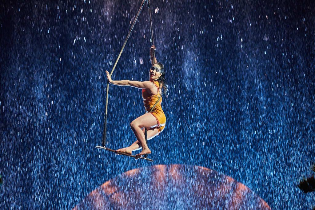 A woman in an orange outfit performs an aerial silk act amidst a backdrop of falling water and a blue stage in Atlanta, Georgia.
