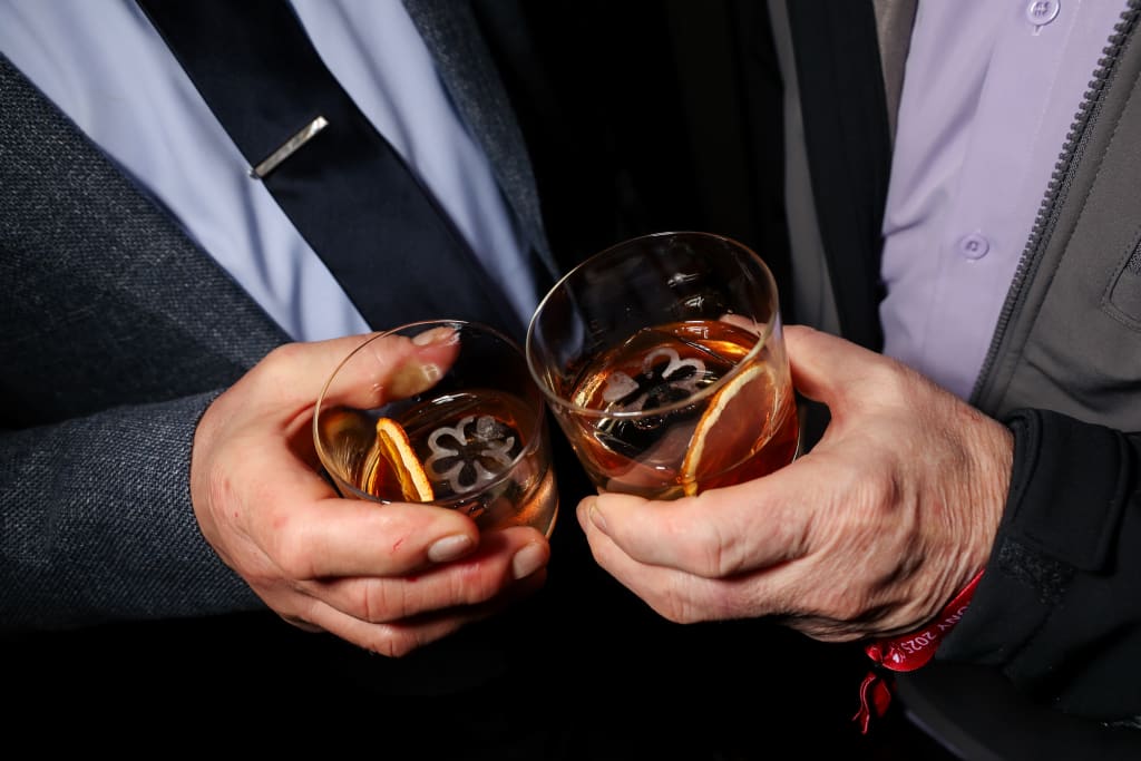 Two men in suits clink glasses filled with amber liquid and ice, garnished with orange slices in Atlanta, Georgia.