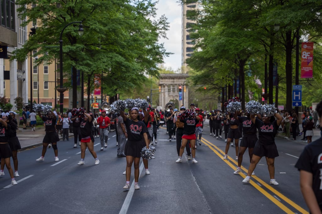 A vibrant parade featuring a marching band and cheerleaders in black and red uniforms, performing on a city street lined with trees and spectators in Atlanta, Georgia.