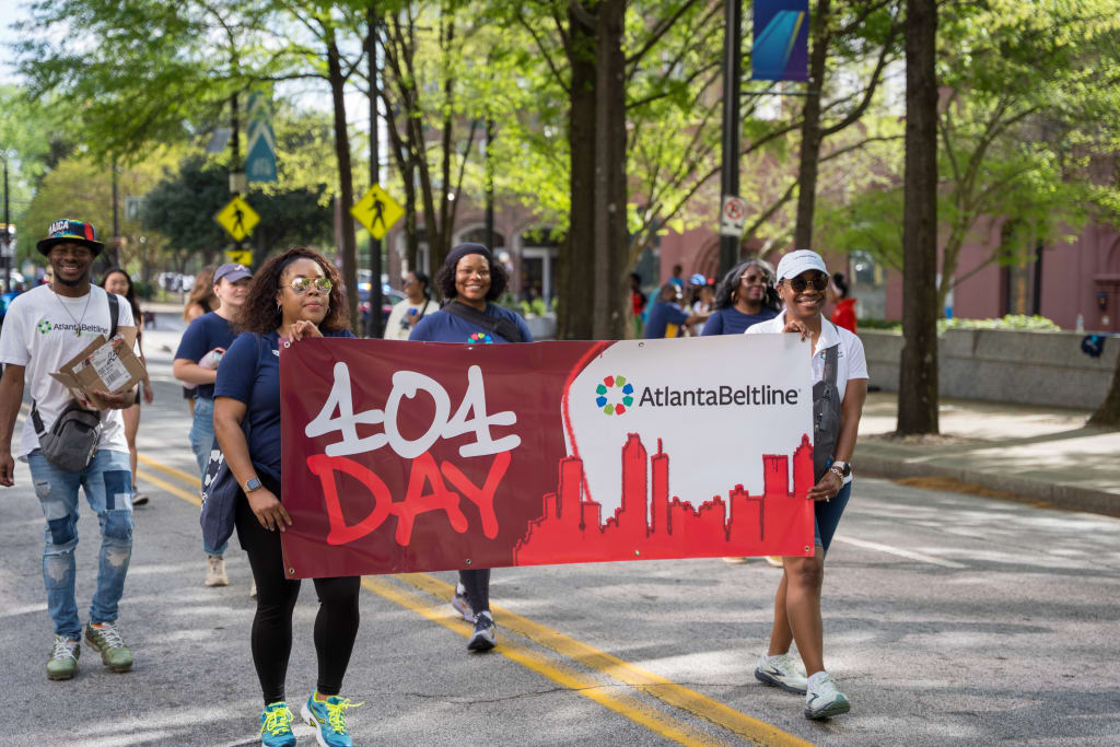 A group of people march down a city street holding a banner that reads '404 Day' with the Atlanta BeltLine logo, surrounded by trees and buildings in Atlanta, Georgia.
