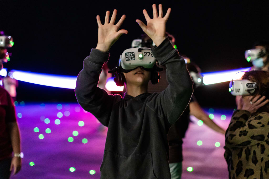 A person wearing a VR headset with their hands raised in excitement, surrounded by others in a dark room illuminated by colorful lights and glowing orbs in Atlanta, Georgia.