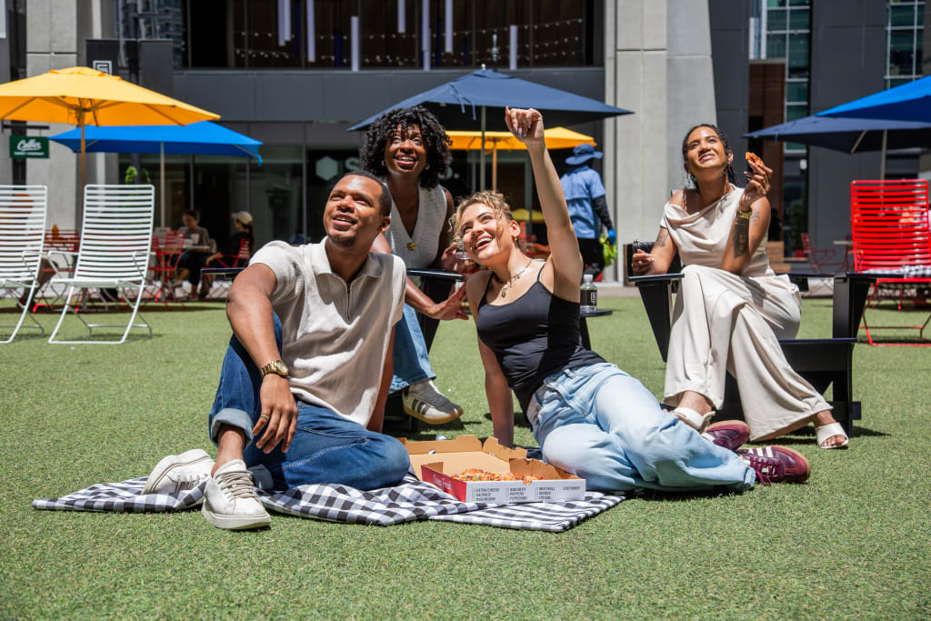 Four friends enjoying a sunny day outdoors, sitting on a checkered blanket with pizza boxes, surrounded by colorful umbrellas and chairs in Atlanta, Georgia.