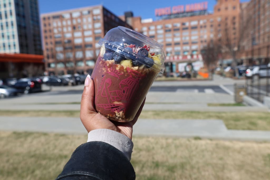 A hand holds a colorful acai bowl topped with berries and granola in a clear plastic cup, with a cityscape featuring the Ponce City Market in the background in Atlanta, Georgia.
