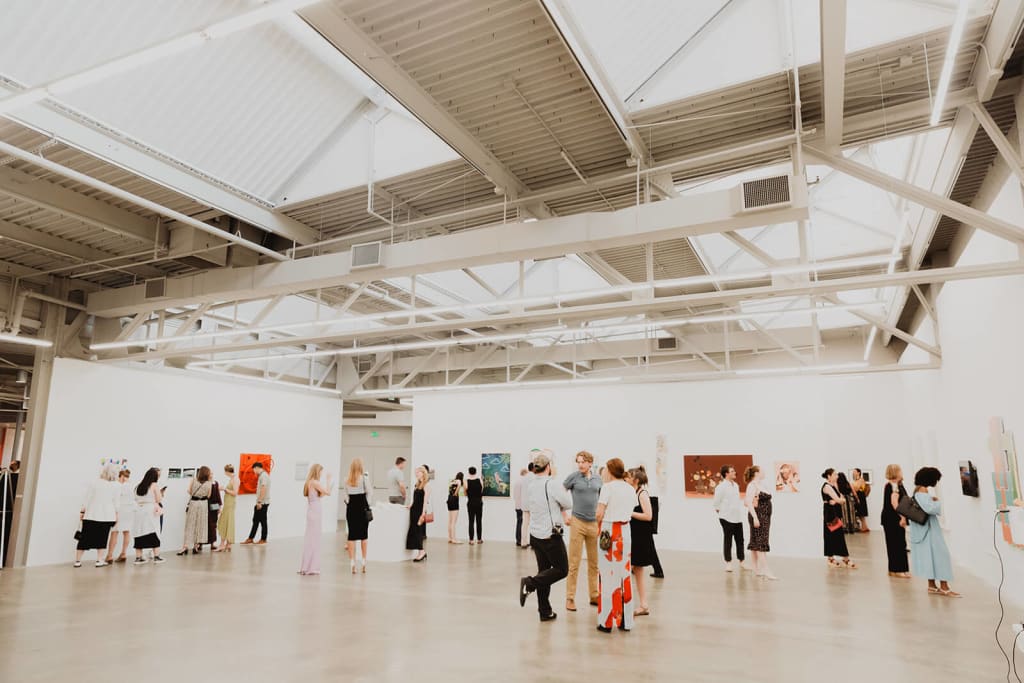 A spacious, well-lit art gallery with high ceilings and white walls, filled with a diverse crowd of people admiring various paintings and artworks in Atlanta, Georgia.