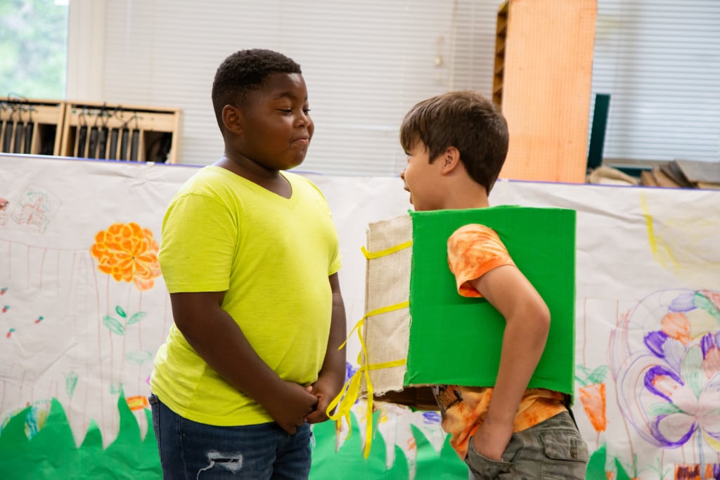 Two young boys, one wearing a green cardboard box costume, engage in a playful conversation in front of a colorful classroom mural in Atlanta, Georgia.