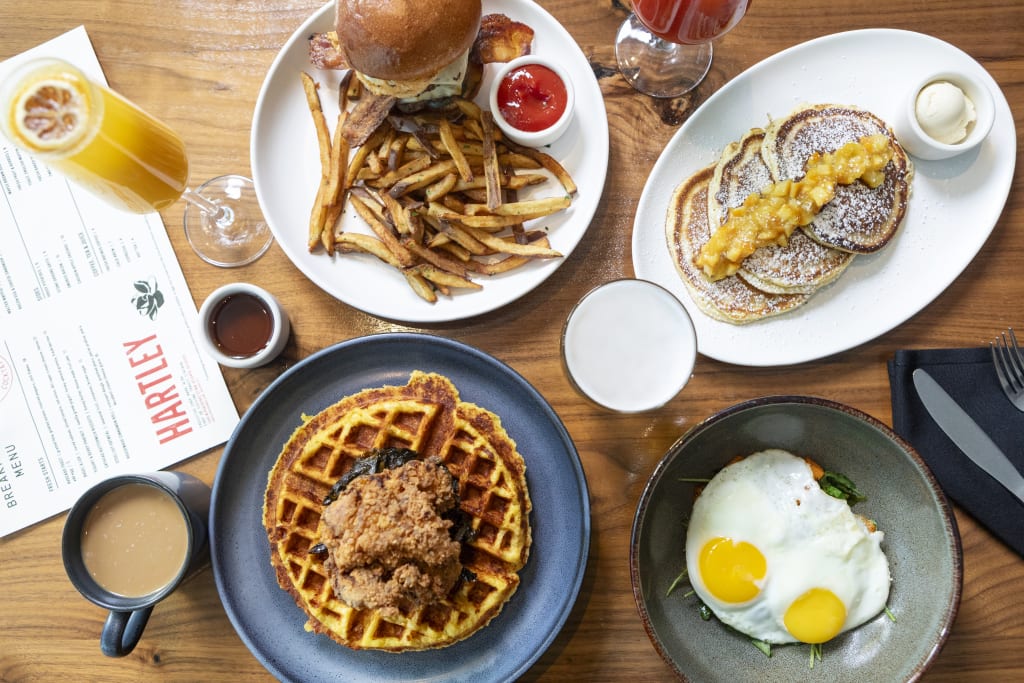 A variety of breakfast and brunch dishes including waffles, pancakes, eggs, bacon, burgers, and fries are spread across a wooden table in Atlanta, Georgia.