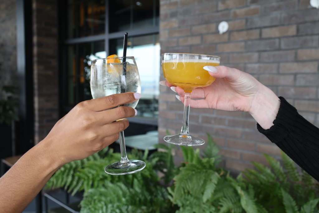 Two hands, one with dark skin and one with light skin, clink glasses filled with a clear beverage and a yellow cocktail, respectively, against a backdrop of lush greenery and a brick wall in Atlanta, Georgia.