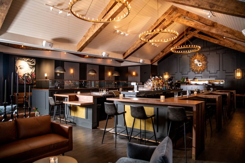 A wide view of the restaurant, Saints and Council, showing the bar, sofas for lounging, and dining spaces. There are two circular chandeliers over the bar. 