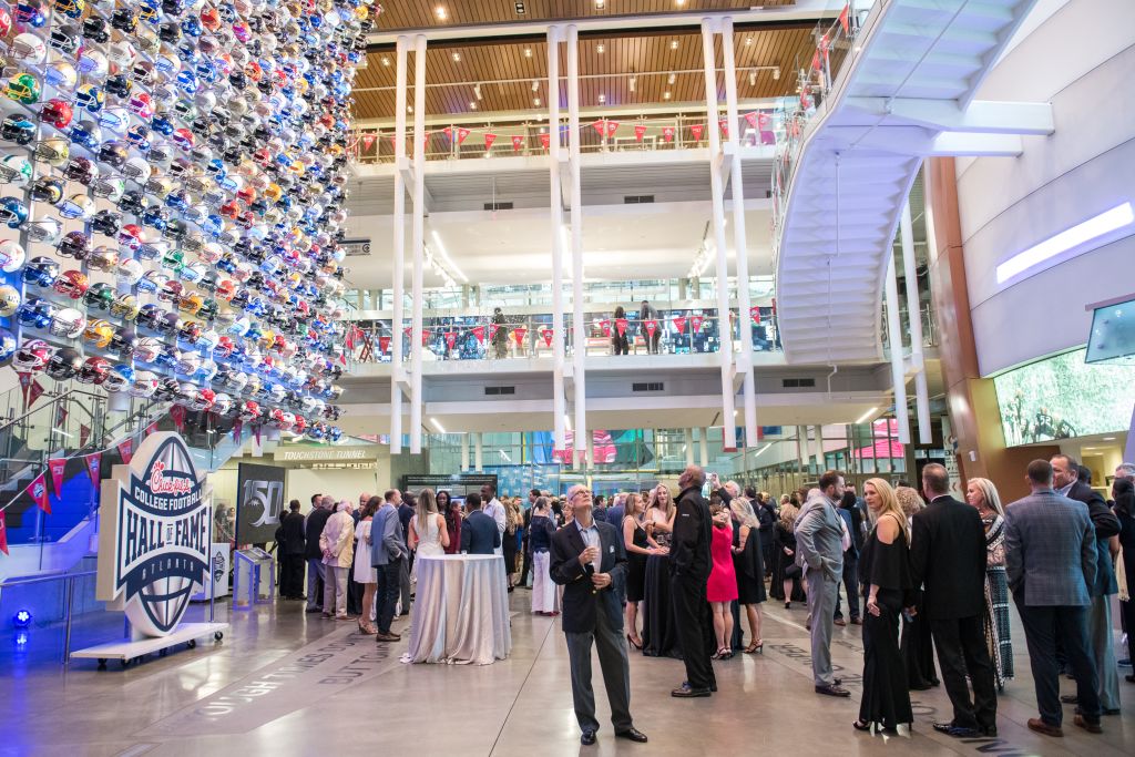 A large crowd gathers in a modern atrium with a football helmet display and a sign for the College Football Hall of Fame in Atlanta, Georgia.