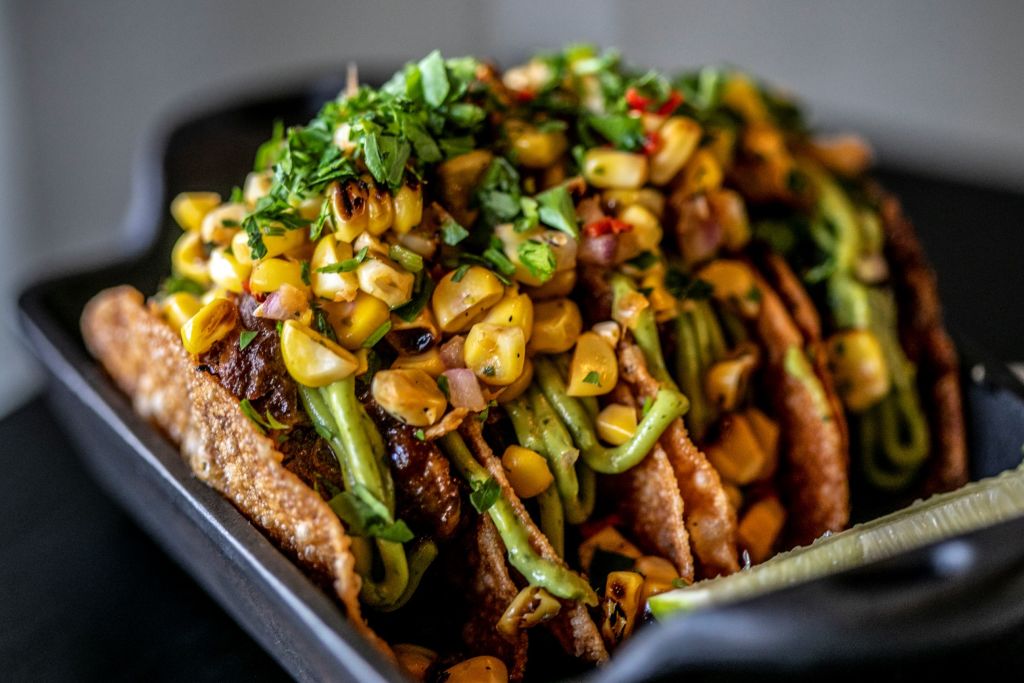 Black plate with 4 tacos in it, covered with corn and cilantro. 