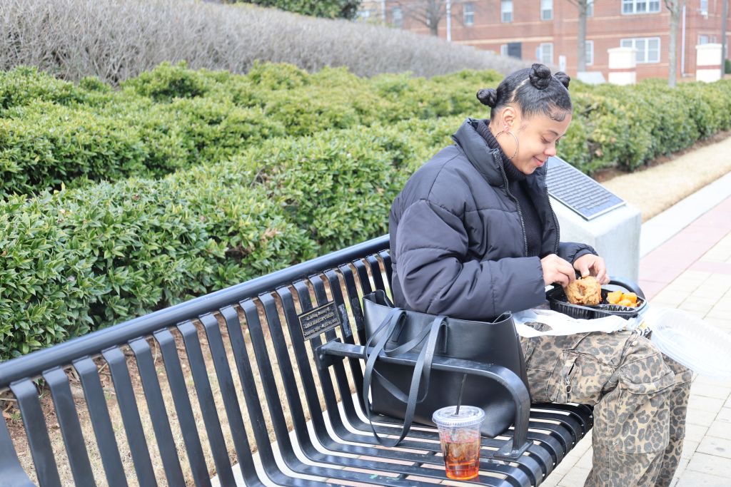 A young woman in a black puffer jacket and leopard print pants sits on a bench eating takeout food in Atlanta, Georgia.