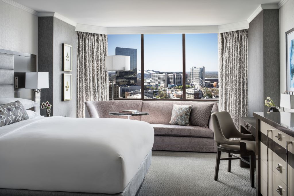 A modern hotel room with a king bed, sofa, desk, and large windows overlooking a cityscape in Atlanta, Georgia.