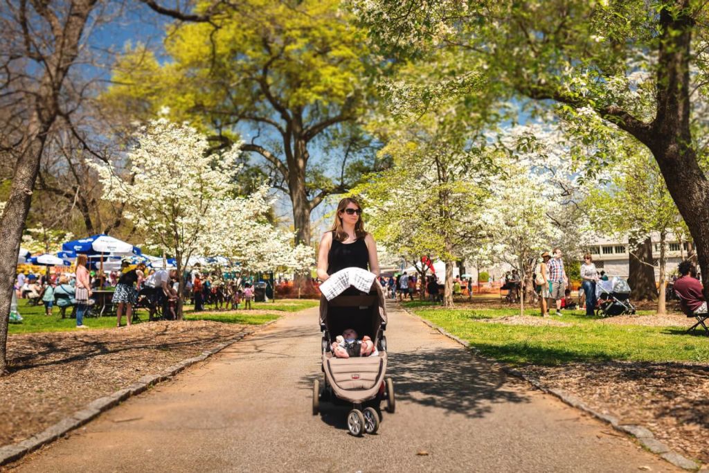 A woman pushes a baby stroller down a tree-lined path in a bustling park on a sunny day in Atlanta, Georgia.