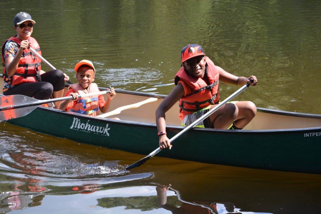 Three people, including two children, are enjoying a sunny day paddling a green canoe on a calm lake, all wearing life jackets and smiling in Atlanta, Georgia.