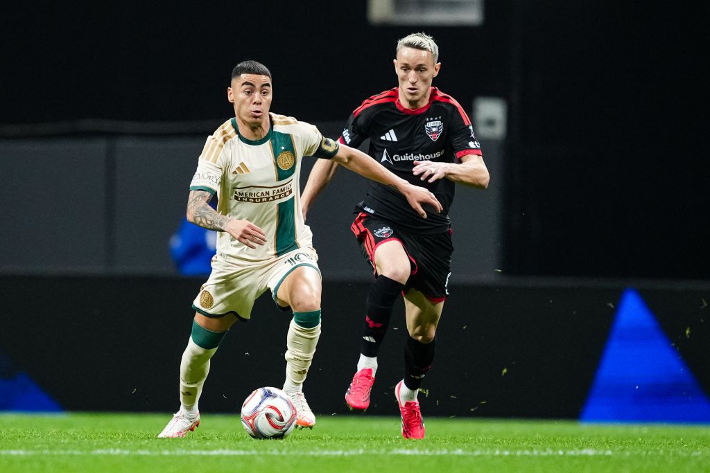 Two soccer players, one in a cream and green uniform and the other in black and red, are sprinting towards a soccer ball on a green field in Atlanta, Georgia.