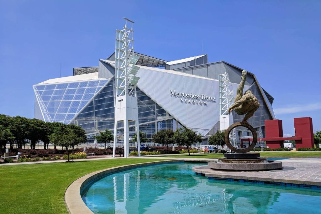 A modern stadium with a distinctive glass facade and a prominent Mercedes-Benz logo, surrounded by a landscaped area with a reflective pool and sculptures in Atlanta, Georgia.