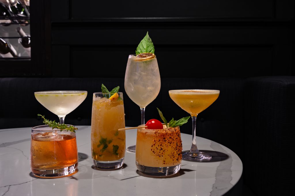 Five vibrant cocktails, each garnished with fresh herbs and fruits, are artfully arranged on a sleek, white marble table against a dark backdrop in Atlanta, Georgia.
