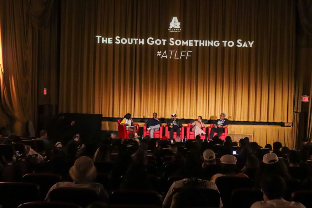 A panel discussion takes place on a stage with a golden curtain backdrop, featuring five individuals seated on red chairs, while an audience fills the theater seats in Atlanta, Georgia.