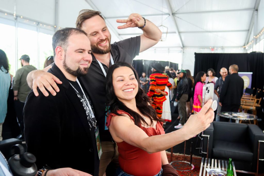 Three friends take a selfie together at a lively event under a large white tent, surrounded by other attendees and colorful decorations in Atlanta, Georgia.