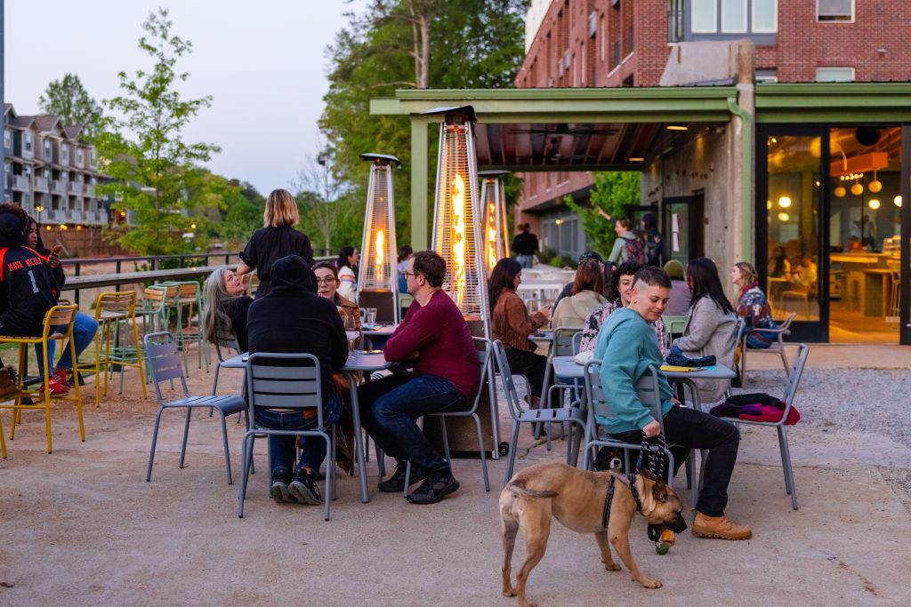 A lively outdoor patio scene at dusk with people dining and socializing around tables with patio heaters, while a dog explores the area in Atlanta, Georgia.