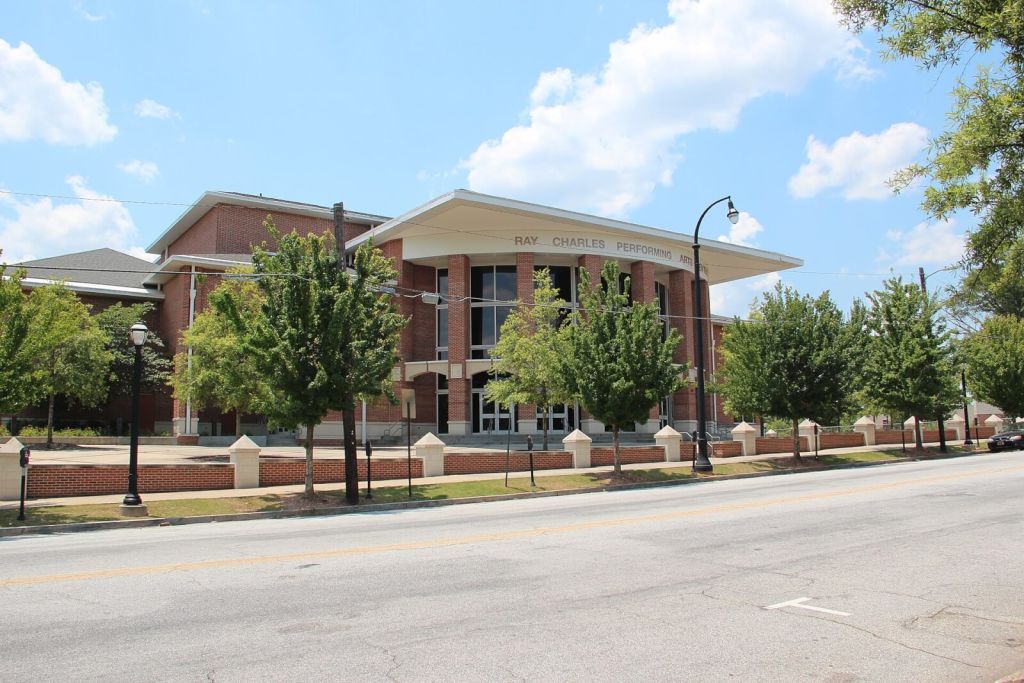 The Ray Charles Performing Arts Center, a modern brick building with large windows and a curved roof, stands prominently on a tree-lined street in Atlanta, Georgia.