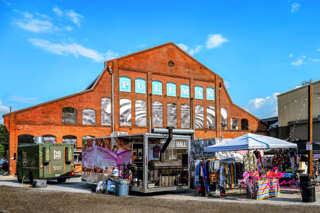 A bustling outdoor market with food trucks, clothing stalls, and a large red brick warehouse in the background in Atlanta, Georgia.