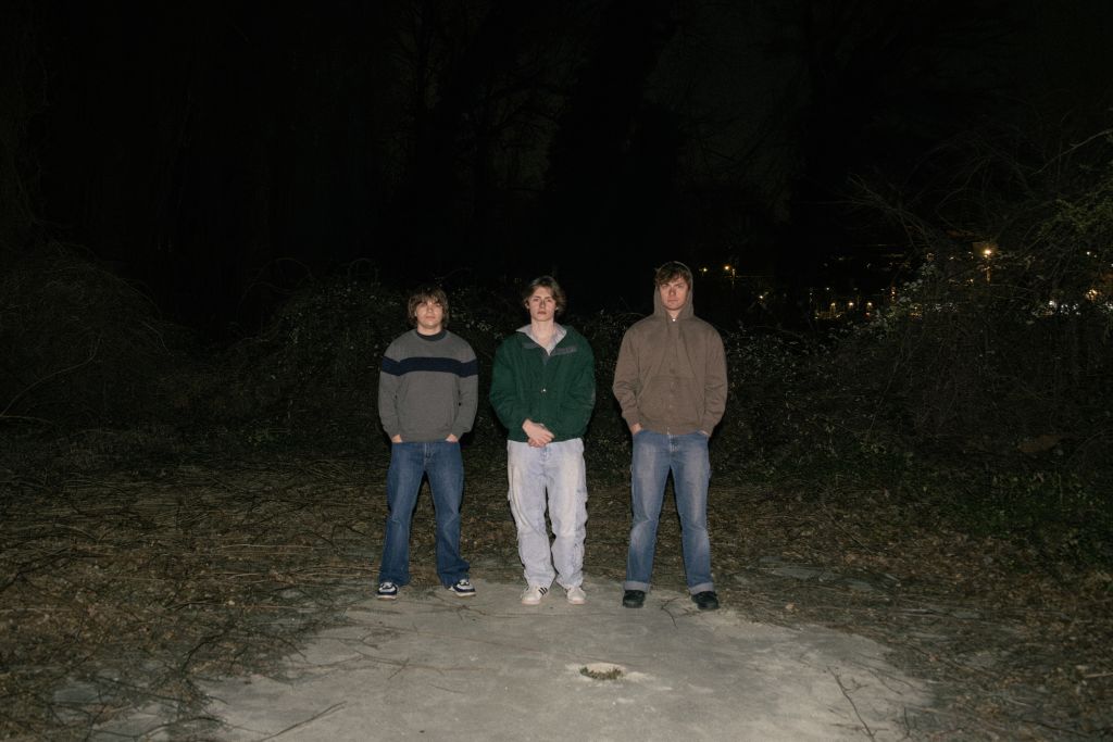 Three young men stand in a line on a dirt path at night, surrounded by dark trees and underbrush in Atlanta, Georgia.