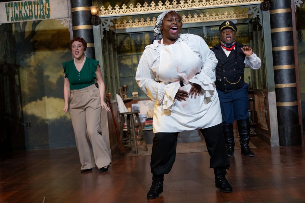 Three actors in period costumes perform a lively scene on a stage set resembling a Victorian-era town, with one actor in a white apron appearing to be in distress while the others react with surprise and concern in Atlanta, Georgia.