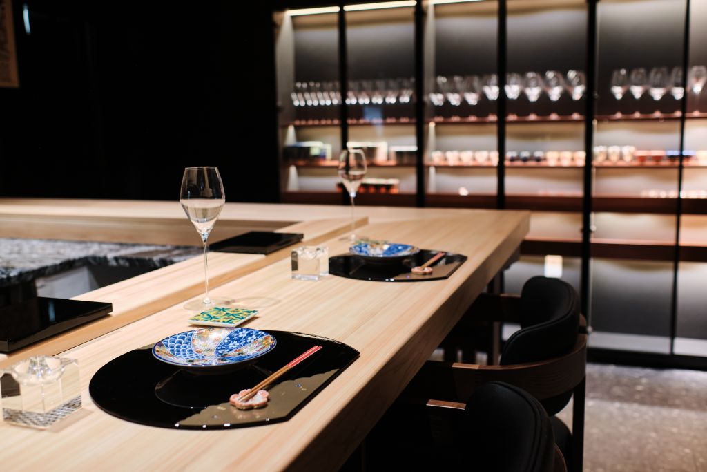 A sleek, modern dining setup with a minimalist wooden table, two blue and white ceramic plates, chopsticks, and wine glasses, all set against a backdrop of illuminated glass shelves filled with various glassware in Atlanta, Georgia.