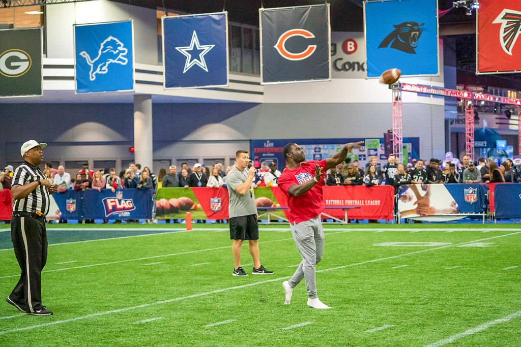 A football player in a red jersey throws a pass during an indoor event, with a referee and crowd watching in Atlanta, Georgia.