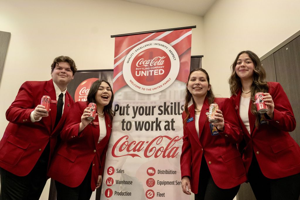 Four individuals in red Coca-Cola uniforms hold cans of soda in front of a banner promoting employment opportunities in Atlanta, Georgia.