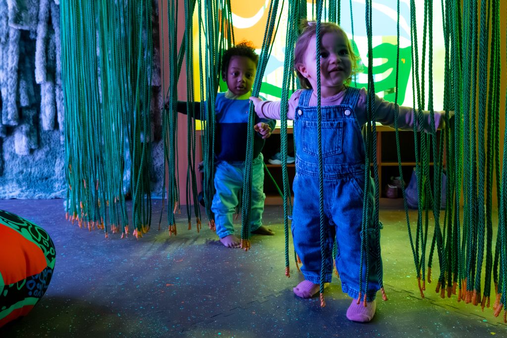 Two young children playfully explore a vibrant, jungle-themed indoor play area with hanging green ropes and colorful lighting in Atlanta, Georgia.