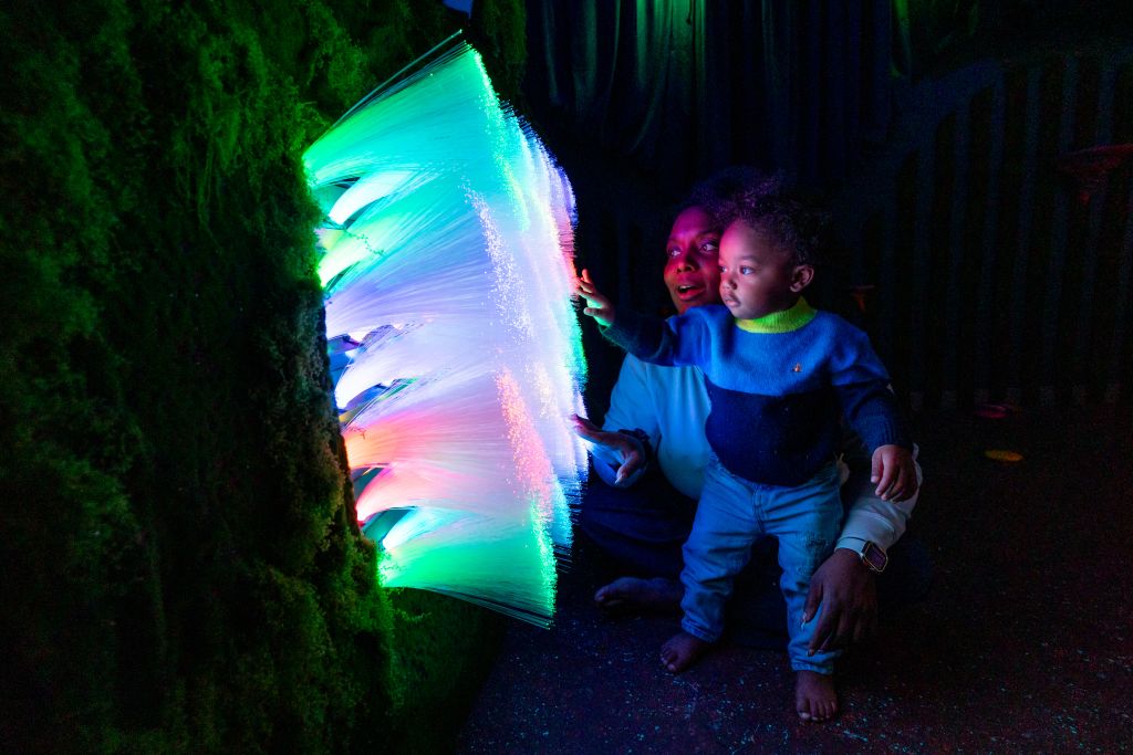 A child and an adult are captivated by a mesmerizing display of colorful, glowing fibers emerging from a dark, moss-covered wall in Atlanta, Georgia.