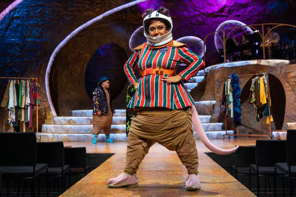 A performer dressed in a colorful, striped space suit with a large helmet and a long tail walks confidently down a runway, surrounded by a fantastical, otherworldly set with glowing walls and floating clothes in Atlanta, Georgia.