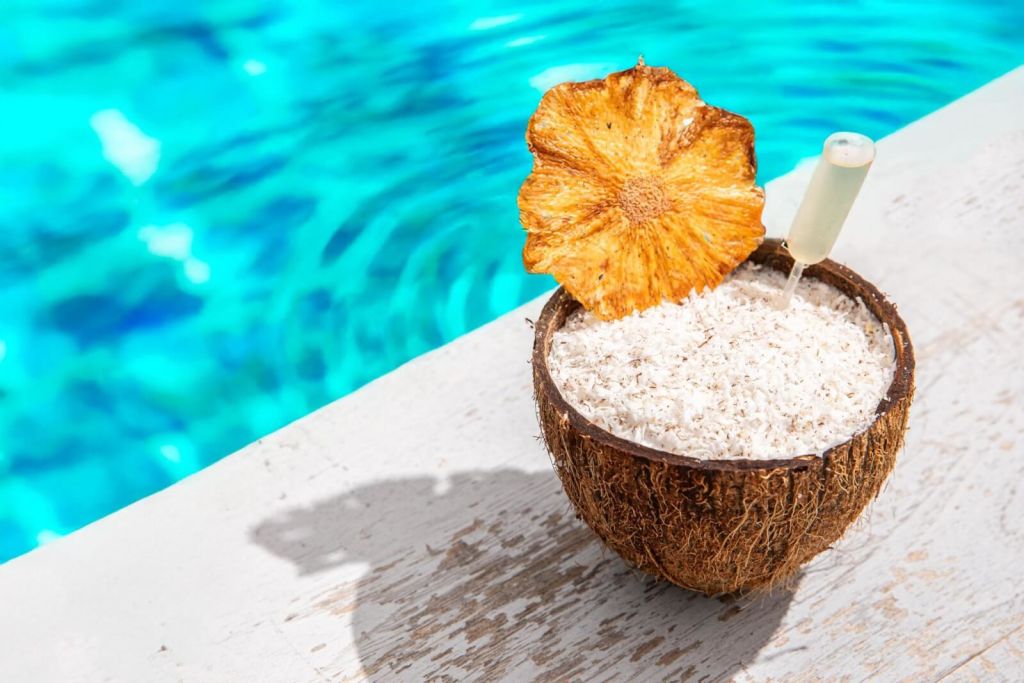 A refreshing coconut cocktail garnished with a dried pineapple slice sits in a coconut shell beside a shimmering blue pool in Atlanta, Georgia.