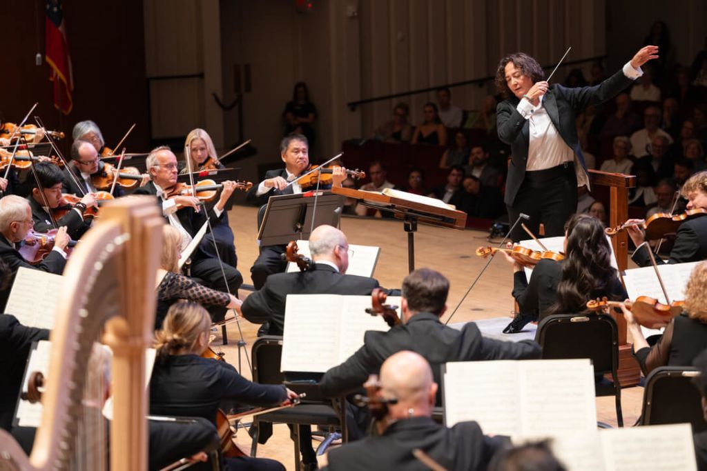A symphony orchestra performs under the direction of a conductor, with musicians playing a variety of instruments including violins, violas, cellos, and a harp in Atlanta, Georgia.