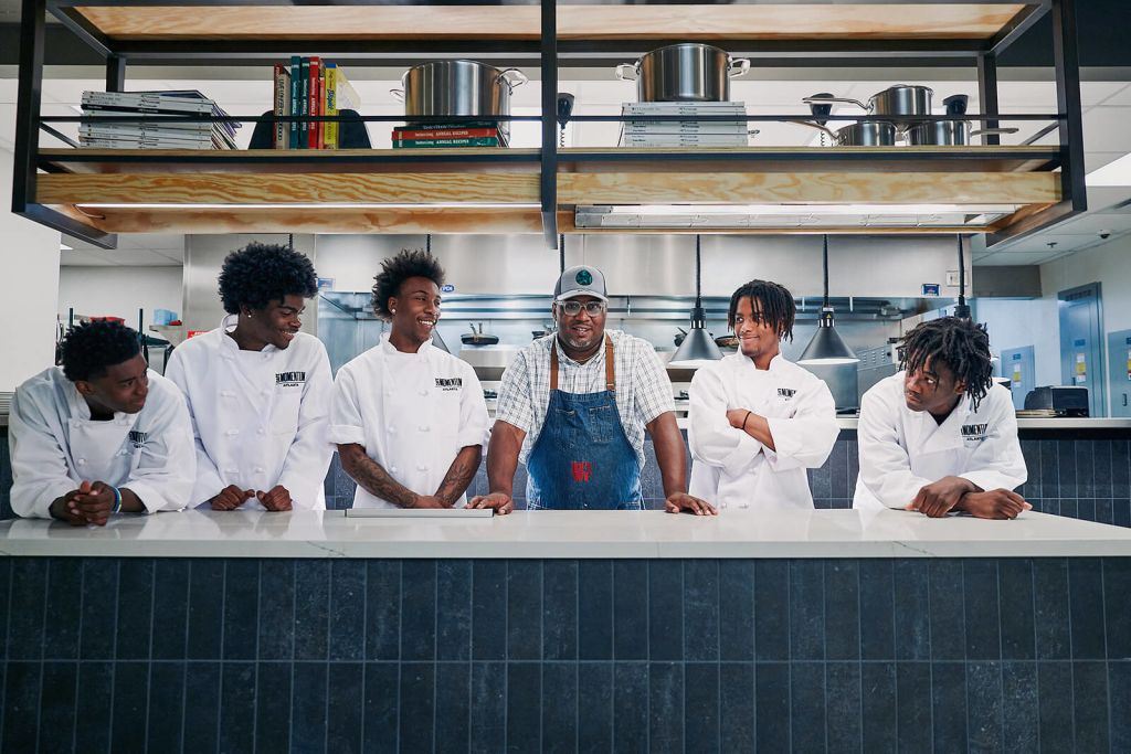 A group of six chefs, five in white coats and one in a denim apron, stand together in a modern kitchen with stainless steel appliances and wooden accents in Atlanta, Georgia.