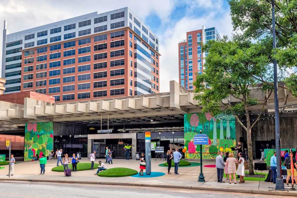 A vibrant urban scene unfolds beneath an elevated highway, where a diverse crowd gathers amidst lush greenery, colorful murals, and modern architecture in Atlanta, Georgia.
