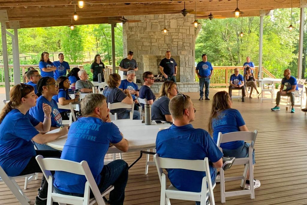 A group of people wearing blue shirts with 'Team Cox Build a Better Future' are gathered on a wooden deck with a stone fireplace, surrounded by lush greenery in Atlanta, Georgia.