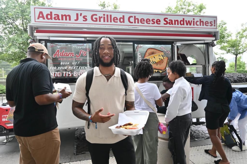 A group of people gather around a food truck named Adam J's Grilled Cheese Sandwiches, enjoying their meals and the sunny day in Atlanta, Georgia.