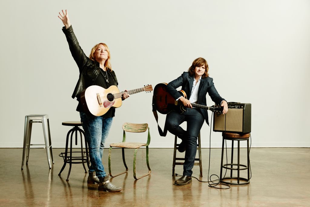Two musicians, one standing with an acoustic guitar and the other seated with an electric guitar, perform in a minimalist studio setting in Atlanta, Georgia.