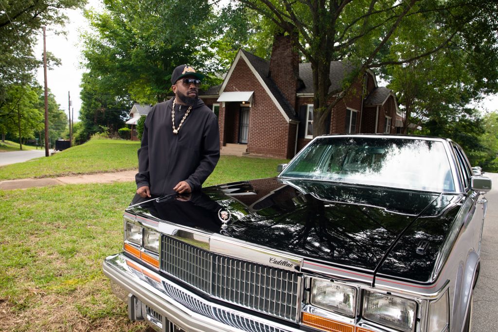 A man in a black hoodie and cap stands next to a classic black Cadillac in front of a brick house with a tree-lined driveway in Atlanta, Georgia.