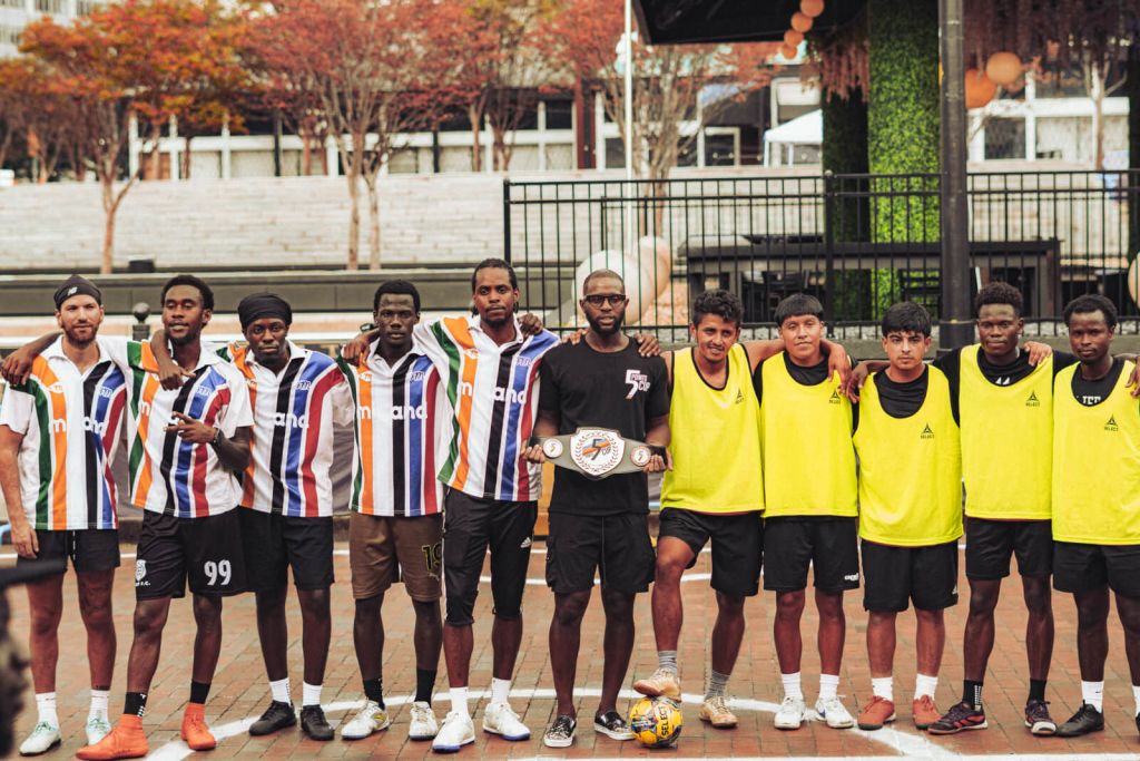A diverse group of young men, dressed in colorful sports jerseys and yellow vests, pose together on a basketball court, holding a championship belt and a soccer ball in Atlanta, Georgia.
