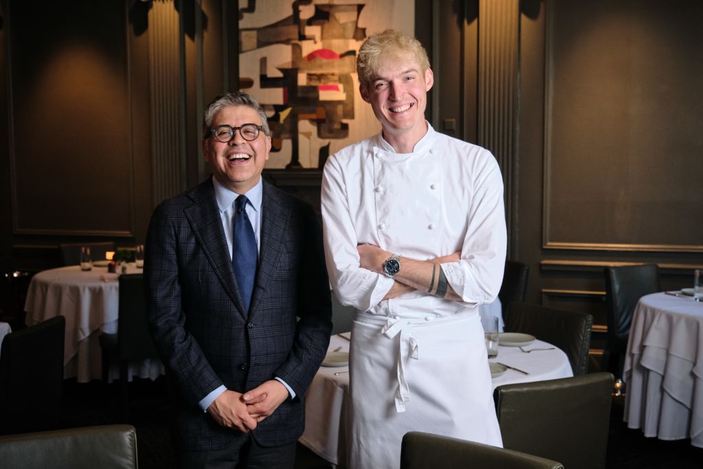 Two men, one in a suit and the other in a chef's uniform, stand together in a restaurant with white tablecloths and elegant decor in Atlanta, Georgia.