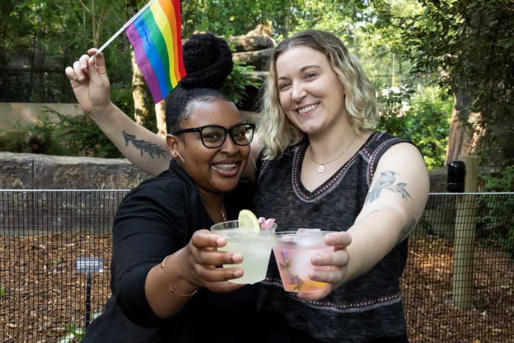 Two women celebrate with rainbow flag and colorful drinks in a garden setting in Atlanta, Georgia.