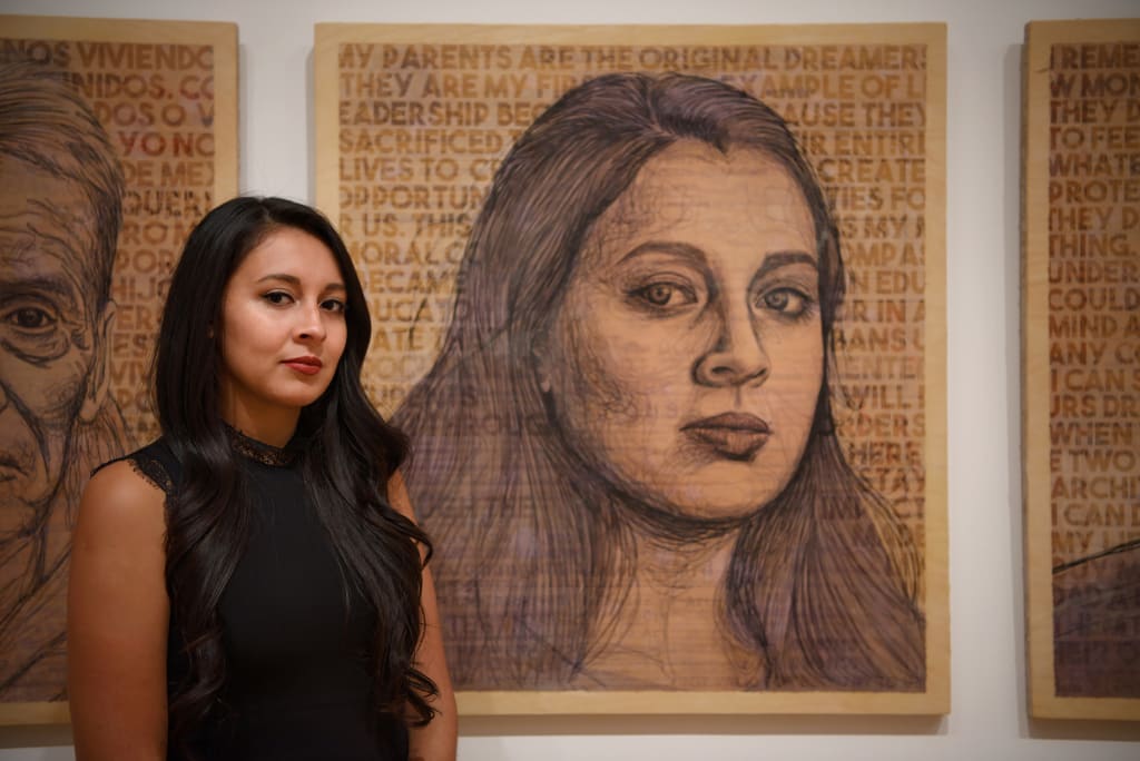 A woman with long dark hair stands in front of three large, detailed pencil portraits on a white wall in Atlanta, Georgia.
